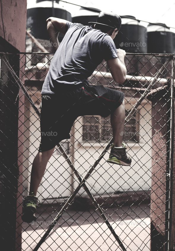 Young man climbing over a chain link fence in an urban environment.