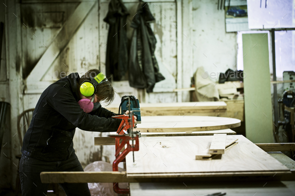 A woman working in a furniture maker\'s workshop cutting timber with a saw.