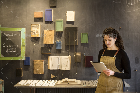 Woman with curly brown hair wearing apron standing in her pottery shop, looking at digital tablet, - Stock Photo - Images