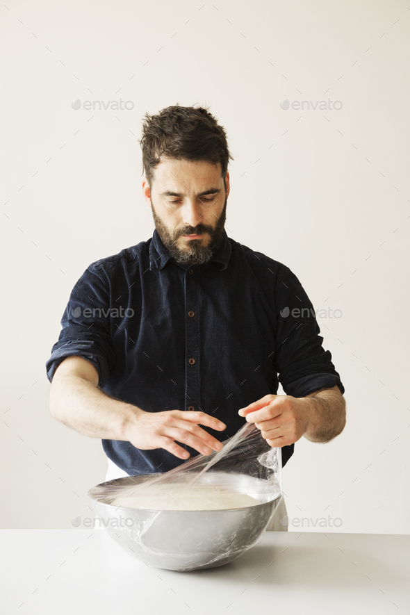 Baker covering bread dough in a metal bowl with cling film.