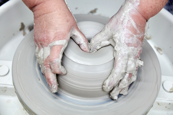 A person using a potter\'s wheel, throwing a clay pot and using thumbs to shape the wet clay. Seen
