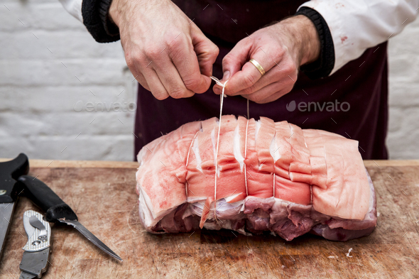 Close up of butcher wearing apron standing at a wooden butcher\'s block, preparing a rolled pork