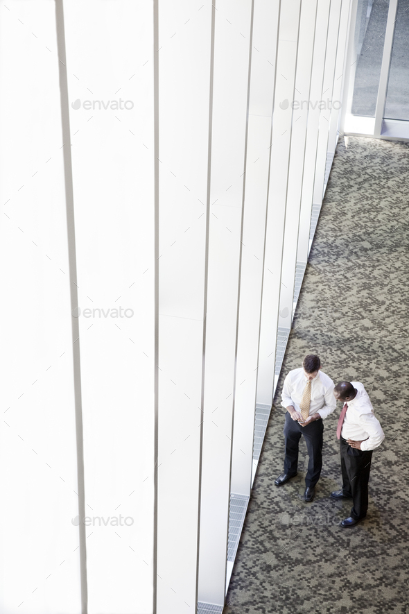 Two businessmen meeting next to a large bank of windows in a business center.