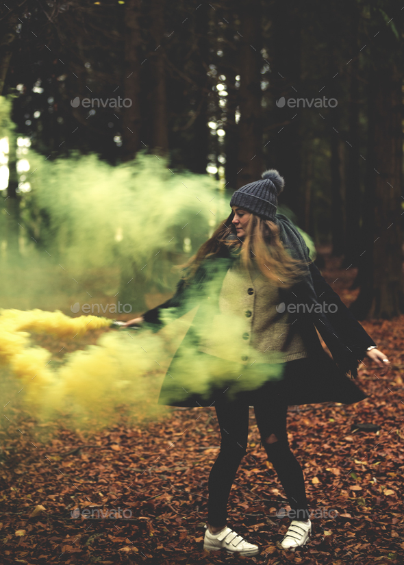 A young woman twirling with a yellow smoke flare in a forest.