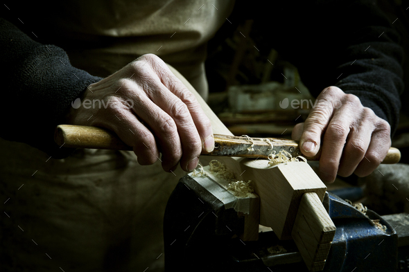 A man working in a furniture maker\'s workshop, using a rasp on a piece of wood in a clamp.