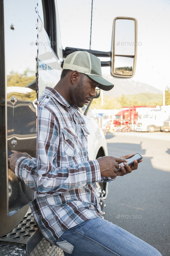 Black man truck driver texting while standing next to his truck cab parked in a lot at a truck stop.
