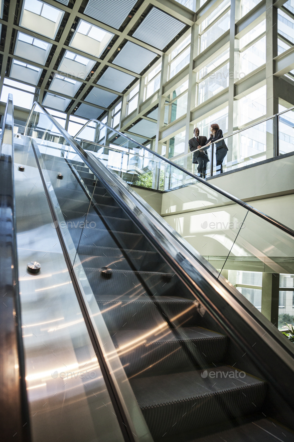 Business man and woman talking at the top of an escalator in a large glassed in open lobby area..