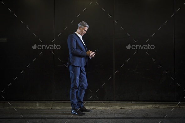 A man in a suit standing in shadow on a city street, looking at his smart phone, texting or - Stock Photo - Images