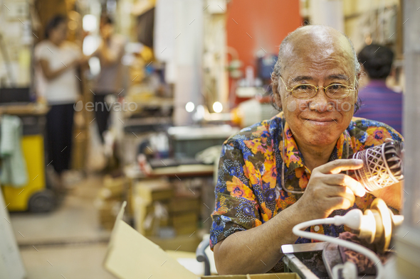 A senior craftsman in a glass maker\'s workshop sitting at a workbench using a light to check an