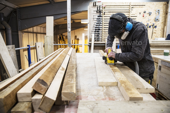Man wearing ear protectors, protective goggles and dust mask standing in a warehouse, sanding planks