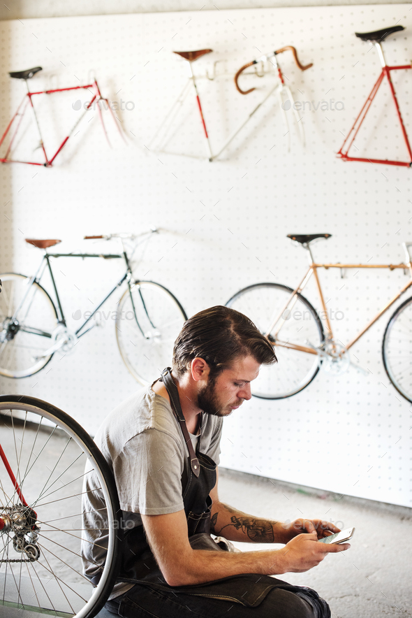 A man working in a bicycle repair shop sitting using his smart phone.