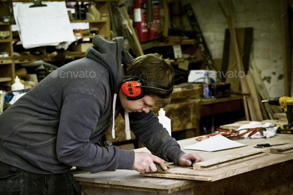 A man working in a furniture maker\'s workshop wearing ear defenders and using a sharp chisel on