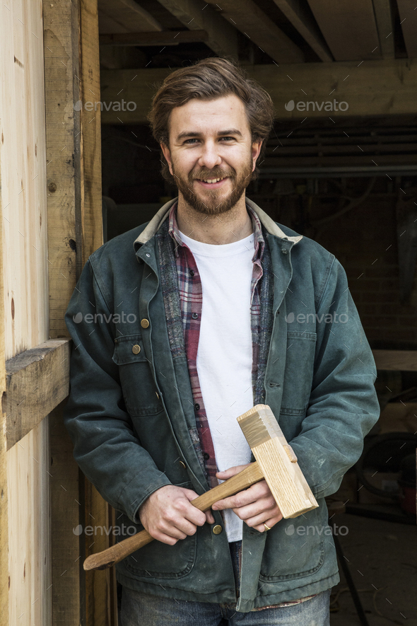 Bearded man standing in doorway of woodworking workshop, holding wooden mallet, smiling at camera.