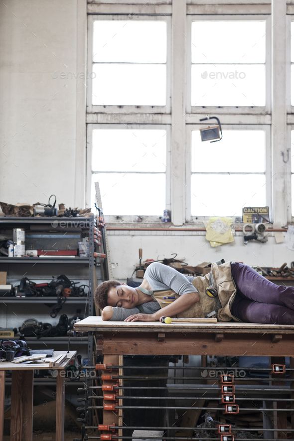 Black woman factory worker taking a nap on top of a work station in a woodworking factory.