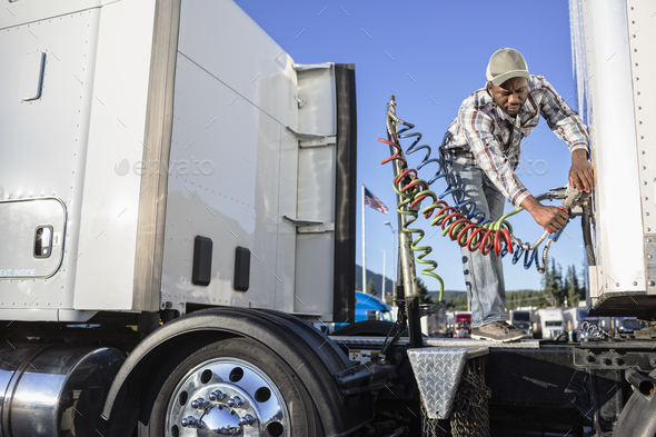 Black man truck driver attaching power cables from truck tractor to trailer at a truck stop.