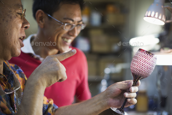 Two people, a father and son at work in a glass maker\'s studio workshop, inspecting a red cut glass