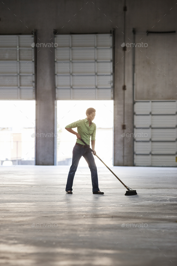 Black woman sweeping up in an empty warehouse space.