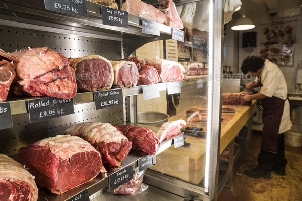Close up of various cuts of beef on shelves in a butcher shop, man wearing apron standing at