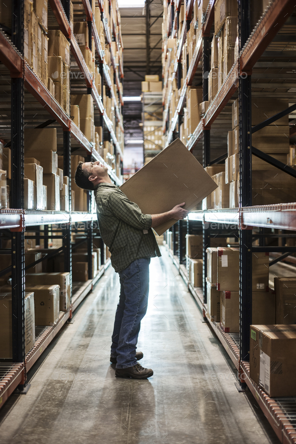 Caucasian male warehouse worker standing in an aisle, holding a box and checking inventory on stacks - Stock Photo - Images