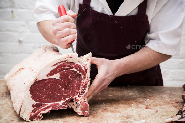Close up of butcher wearing apron standing at a wooden butcher\'s block, butchering beef forerib.