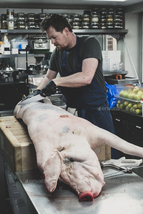 Male butcher wearing apron and black rubber gloves cutting pig\'s carcass on butcher\'s block.