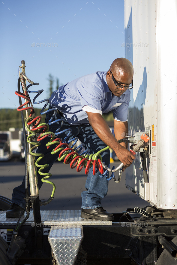 Black man truck driver attaching power cables from truck tractor to trailer at a truck stop.
