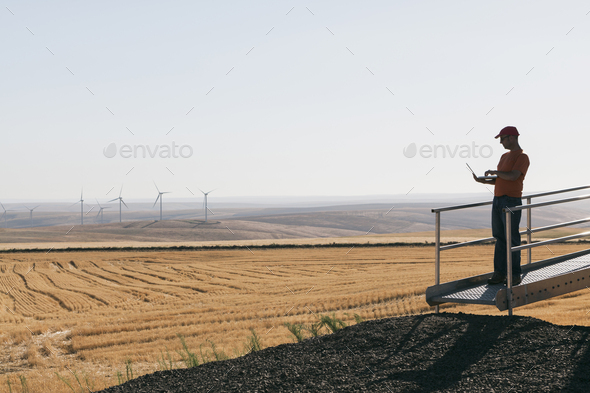 A wind farm technician standing and using a laptop at the base of a turbine on a wind farm in open