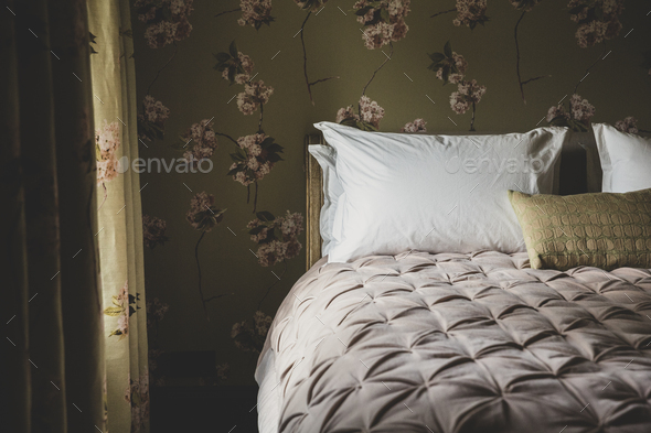 Interior view of bedroom with curtains and wallpaper with floral pattern, pale pink quilt and white