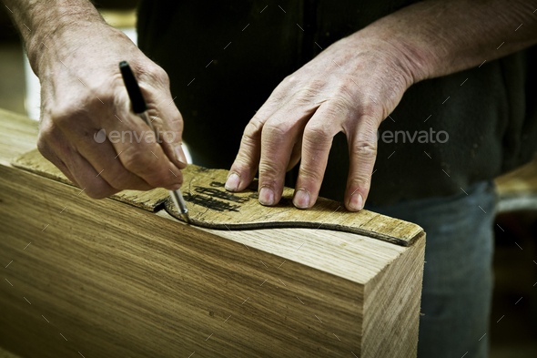A man working in a furniture maker\'s workshop drawing around a shape on wood.
