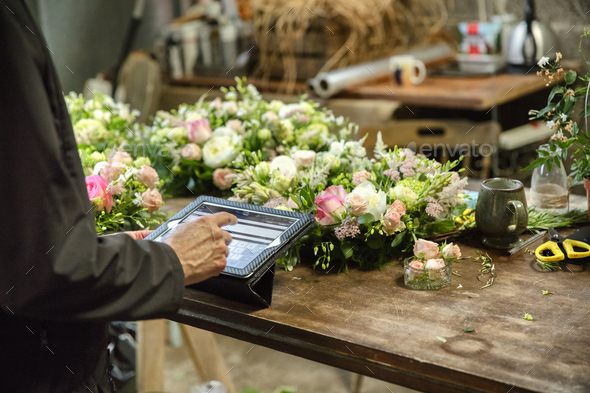 A woman in a florist's workshop using a digital tablet, at a workbench with flower arrangements.