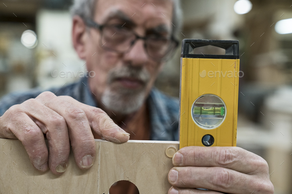 A senior man with glasses and beard in a woodworkers shop, using a spirit level checking his work.