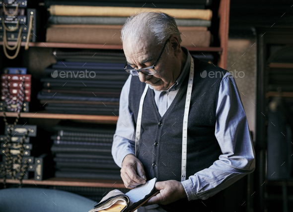 Tailor with measuring tape around his neck inspecting fabric samples.