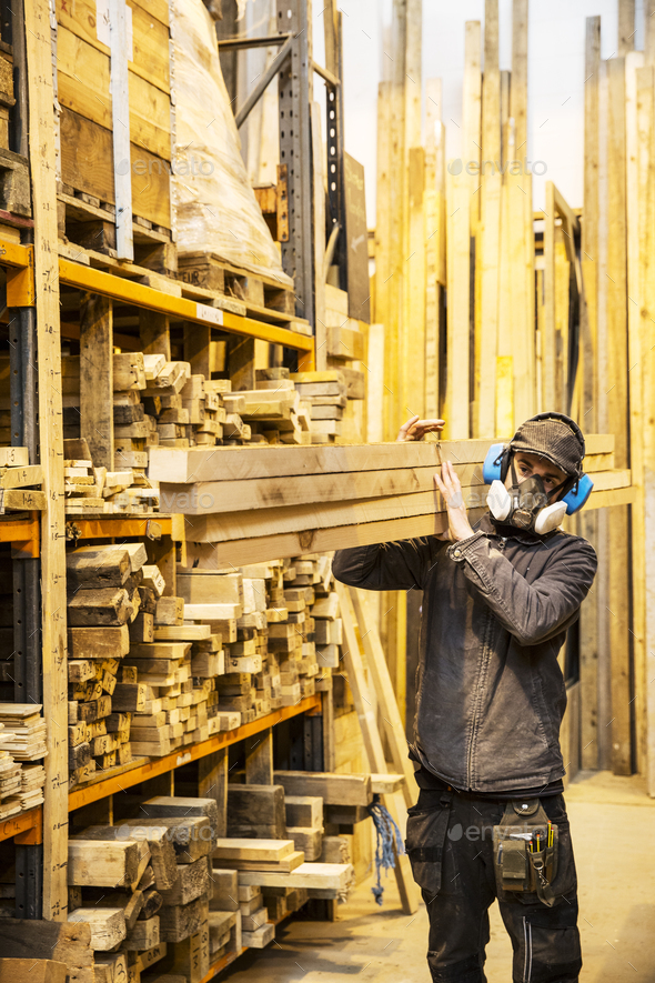 Man wearing ear protectors and dust mask standing in a warehouse, carrying long planks of wood on