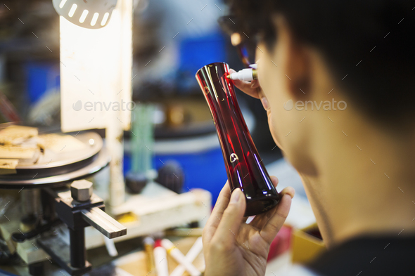 A craftsman in a glass maker\'s workshop inspecting a red glass vase and marking the exterior.