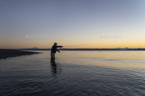 A silhouette view of a fly fisherman casting for salmon and searun coastal cutthroat trout from a - Stock Photo - Images
