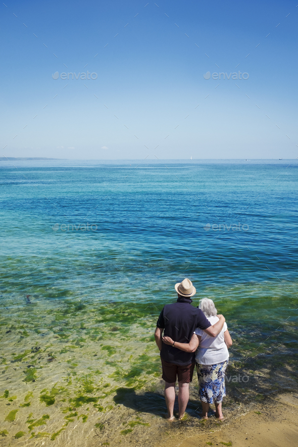 Rear view of elderly couple standing with arms around shoulders looking out to sea across the vivid