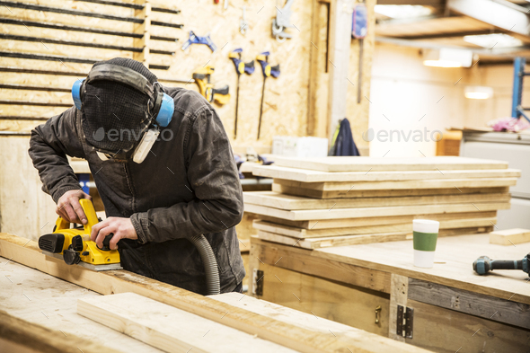 Man wearing ear protectors, protective goggles and dust mask standing in a warehouse, sanding planks