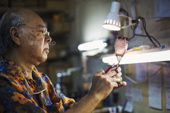 A senior craftsman at work in a glass maker's studio workshop, in inspecting red wine glass with cut - Stock Photo - Images