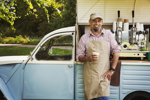 Download Bearded man wearing apron standing by blue mobile coffee shop, holding hot drink, smiling at ...