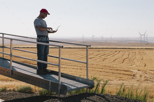 A wind farm technician standing and using a laptop at the base of a turbine on a wind farm in open