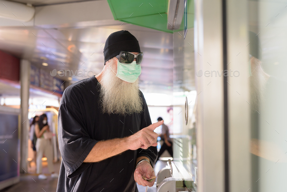 Mature bearded hipster man with mask and sunglasses buying ticket at the sky train station - Stock Photo - Images