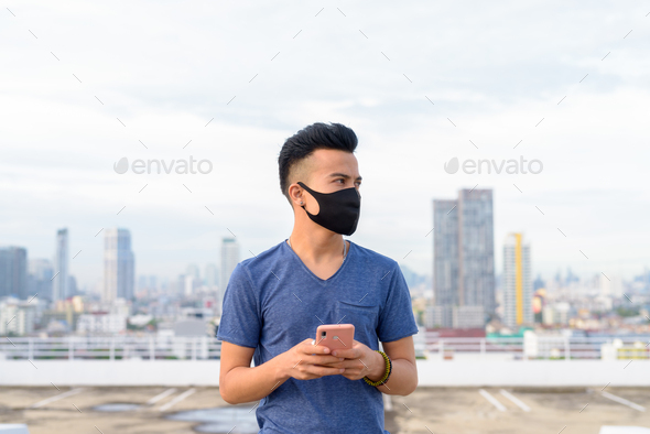 Young multi ethnic man with mask thinking while using phone against view of the city