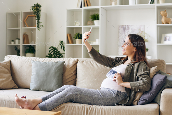 Happy pregnant woman with ultrasonic picture of her baby making selfie on couch - Stock Photo - Images