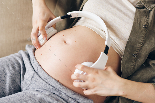 Hands of pregnant female holding white headphones on both sides of her belly