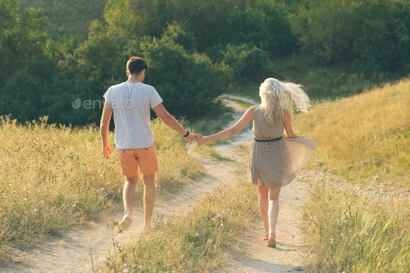 Young couple of man and woman together, hand by hand, are walking down by the field road in summer.
