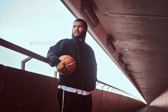 Dark-skinned guy holds a basketball while standing on a footway under bridge. - Stock Photo - Images