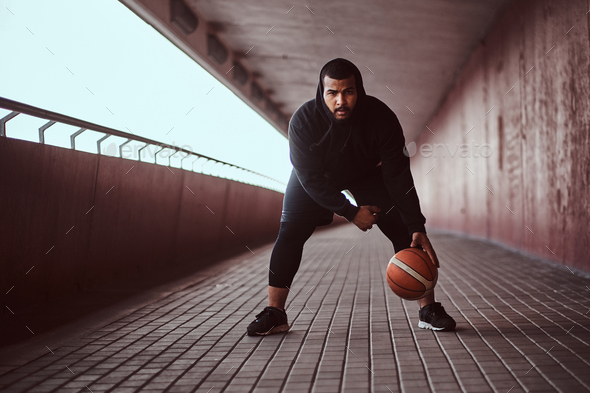 Dark-skinned guy holds a basketball while standing on a footway under bridge. - Stock Photo - Images