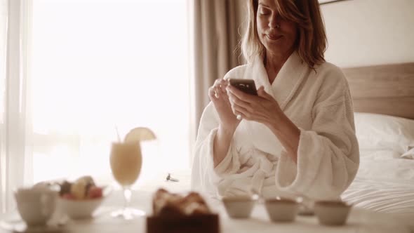Senior woman using smartphone, sitting with breakfast in hotel room alt