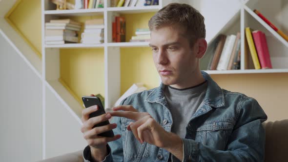 Smiling Young Man Holds Mobile Phone While Sitting on Sofa at Home and Celebrating alt