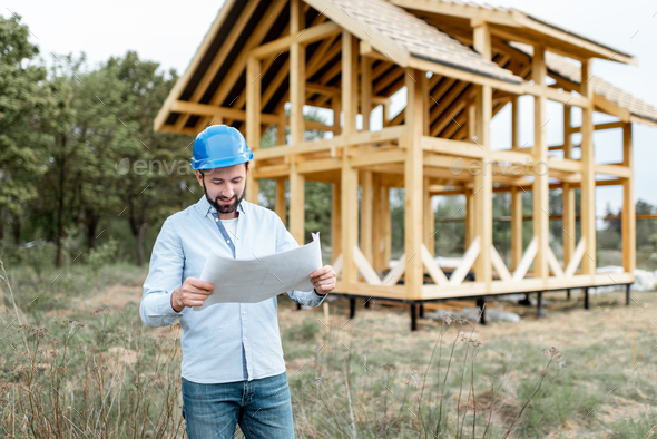 Builder with blueprints near the wooden house on the construction site - Stock Photo - Images
